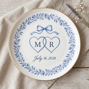 May include: White ceramic plate with a gold rim, featuring a blue floral wreath design. Two intertwined blue hearts with the letters "M" and "R" are centered, topped with a blue bow. The date "July 16, 2026" is written below.