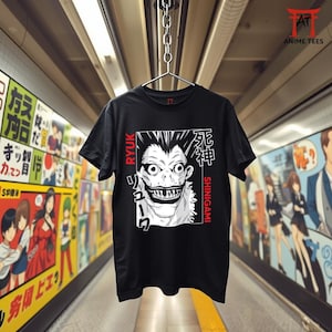 Könnte beinhalten: Schwarzes T-Shirt mit einer weiß-schwarzen Grafik eines Charakters mit scharfen Zähnen und dem Text "RYUK" und "SHINIGAMI" in Rot. Das T-Shirt hängt an einem Metallbügel.