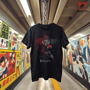 May include: Black t-shirt with a red and black graphic of a character holding a red apple, and the white text "DEATH NOTE". The t-shirt is suspended from a metal hanger.