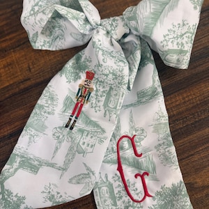 Puede incluir: Lazo con estampado blanco y verde, con un cascanueces y la letra "G" bordada en rojo. El lazo presenta un diseño de toile con edificios y árboles. El lazo está anudado, con los extremos colgando.