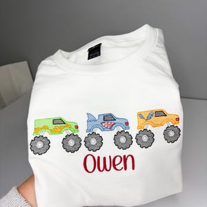 K&ouml;nnte beinhalten: Wei&szlig;es T-Shirt mit gestickten Monstertrucks in Gr&uuml;n, Blau und Orange mit schwarzen R&auml;dern. Der Name "Owen" ist rot unter den Trucks gestickt. Das Shirt ist gefaltet.