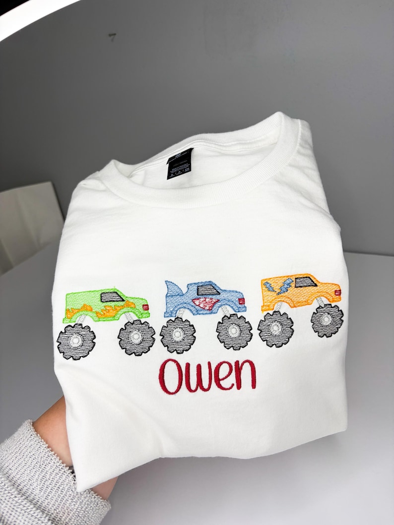K&ouml;nnte beinhalten: Wei&szlig;es T-Shirt mit gestickten Monstertrucks in Gr&uuml;n, Blau und Orange mit schwarzen R&auml;dern. Der Name "Owen" ist rot unter den Trucks gestickt. Das Shirt ist gefaltet.