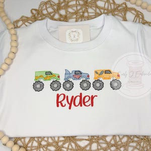 K&ouml;nnte beinhalten: Wei&szlig;es T-Shirt mit gestickten Monstertrucks in Gr&uuml;n, Blau und Gelb mit schwarzen R&auml;dern. Der Name "Ryder" ist rot darunter gestickt. Ein verspieltes Design f&uuml;r Kinder.