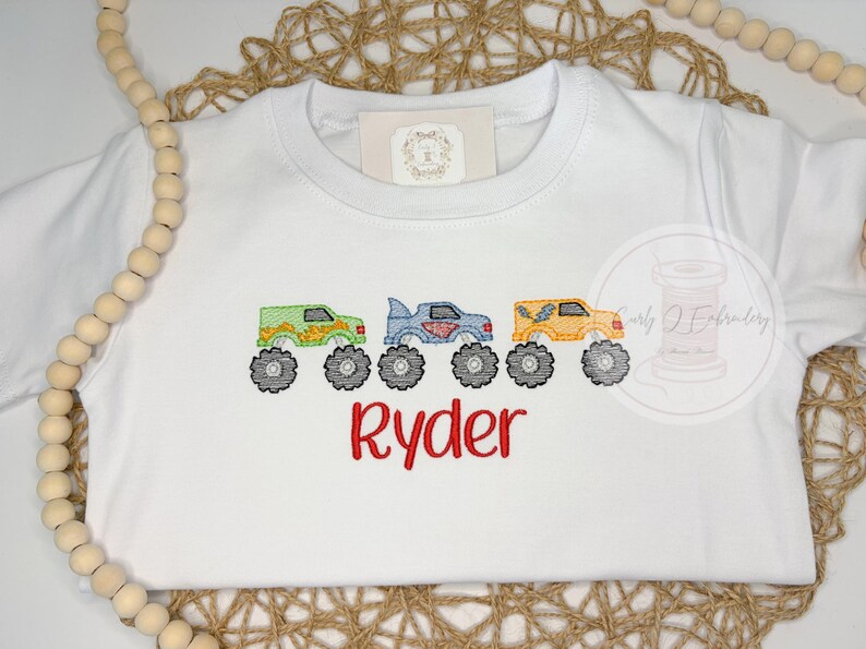 K&ouml;nnte beinhalten: Wei&szlig;es T-Shirt mit gestickten Monstertrucks in Gr&uuml;n, Blau und Gelb mit schwarzen R&auml;dern. Der Name "Ryder" ist rot darunter gestickt. Ein verspieltes Design f&uuml;r Kinder.