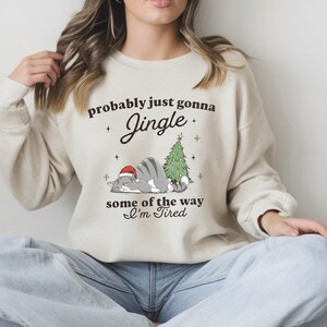 Może przedstawiać: Beżowa bluza z napisem "probably just gonna Jingle some of the way I'm Tired". Grafika przedstawia szarego kota w czapce Świętego Mikołaja, choinkę i gwiazdki. Bluza ma długie rękawy i okrągły dekolt.