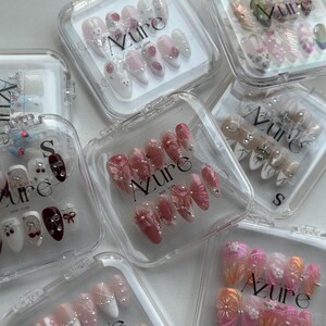 May include: A collection of press-on nails in clear plastic cases. The nails feature various designs, including floral patterns, pearls, and cartoon characters. The brand name "Azure" is visible on the cases. The nails are in various shades of pink, white, and orange.