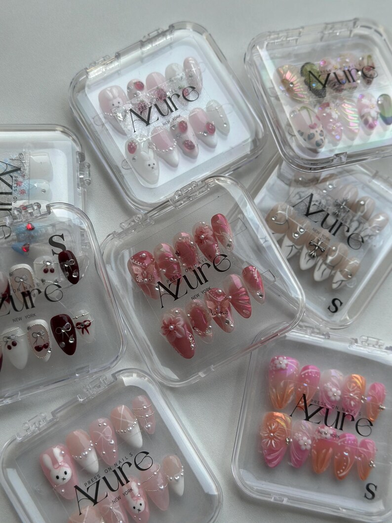 May include: A collection of press-on nails in clear plastic cases. The nails feature various designs, including floral patterns, pearls, and cartoon characters. The brand name "Azure" is visible on the cases. The nails are in various shades of pink, white, and orange.
