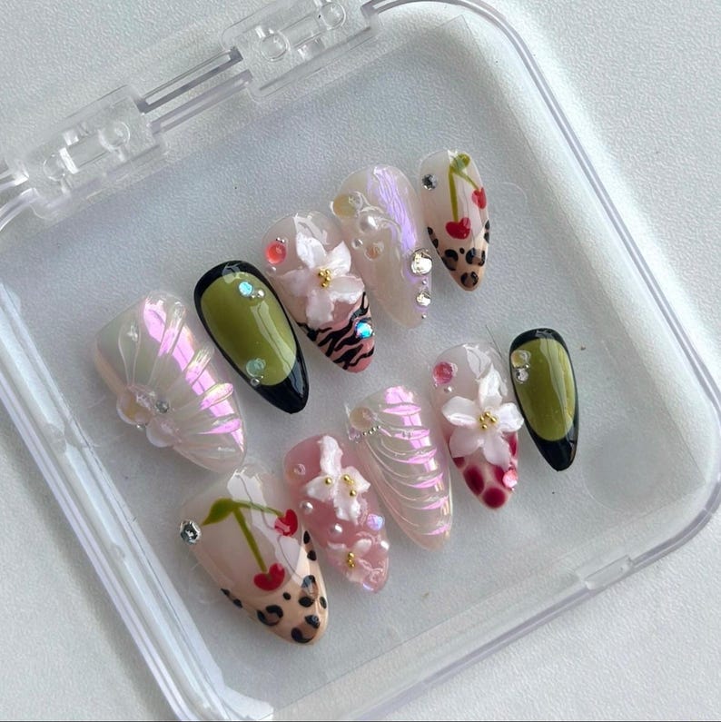 May include: A set of artificial nails in a clear plastic case. The nails feature various designs, including floral accents, cherry motifs, and animal prints. Colors include pink, white, green, and black, with iridescent and glitter details.