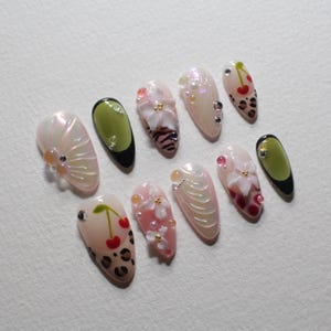 May include: A collection of ten almond-shaped artificial nails with various designs. Some nails feature cherry and leopard print patterns, while others showcase floral and pearl embellishments. The color palette includes pink, white, and green.