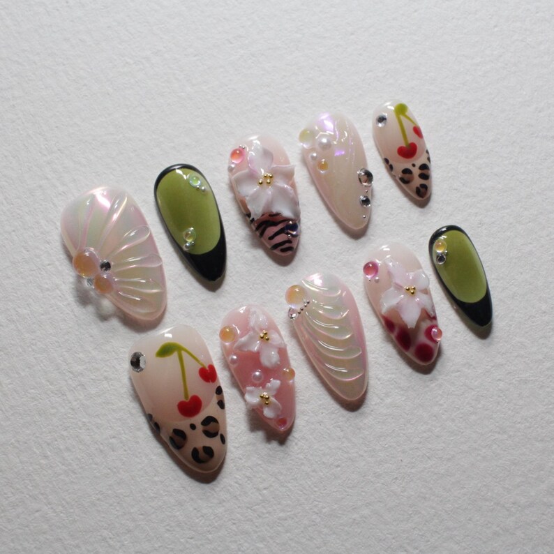 May include: A collection of ten almond-shaped artificial nails with various designs. Some nails feature cherry and leopard print patterns, while others showcase floral and pearl embellishments. The color palette includes pink, white, and green.
