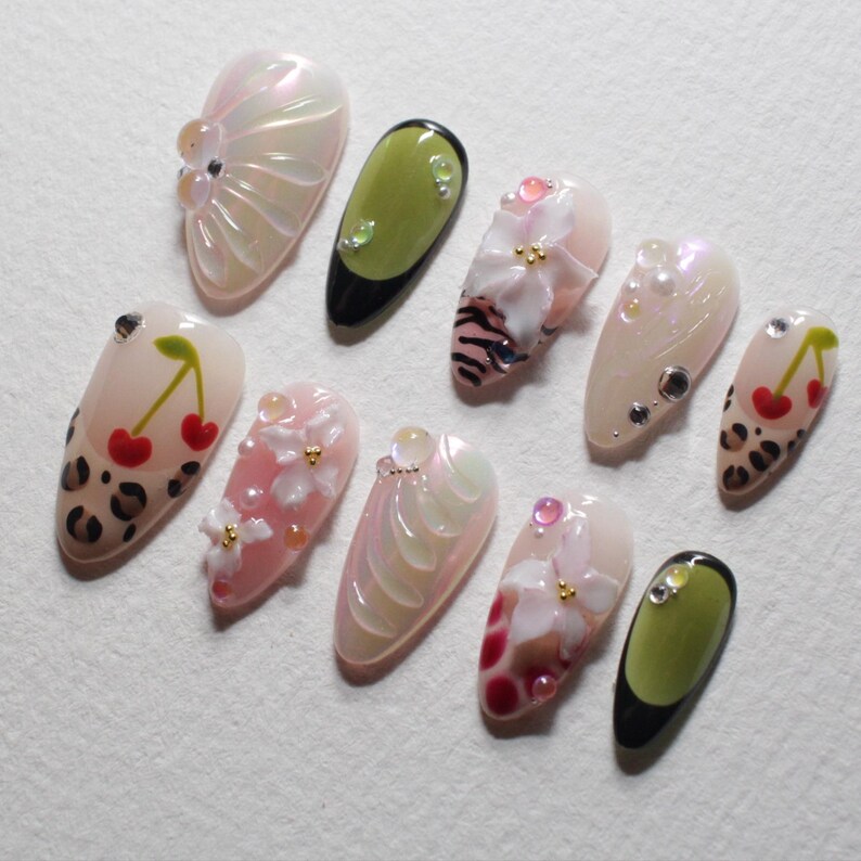 May include: A collection of ten artificial nails with various designs. Some nails feature cherry and leopard print patterns, while others showcase floral and abstract designs with iridescent and pearl accents. The nails are in various shades of pink, white, and green.