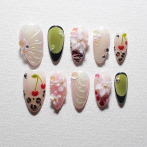 May include: A set of ten artificial nails with various designs. The nails feature a range of colors, including pink, green, and white, with embellishments like cherries, flowers, and glitter. Some nails have a leopard print design.