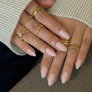 Könnte beinhalten: Nahaufnahme von Händen mit mandelförmigen, hellrosa Nägeln. Mehrere goldene Ringe in verschiedenen Designs schmücken die Finger, darunter ein gestepptes Band, eine Kette und ein Ring mit einem zentralen Stein. Die Nägel sind gepflegt.