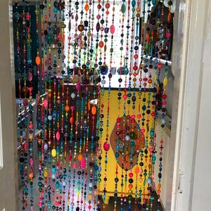 Pu&ograve; includere: Una tenda per porta fatta di fili di perline colorate. Le perline sono di varie forme e dimensioni, tra cui rotonde, ovali e oblunghe, nelle tonalit&agrave; di rosso, arancione, giallo, verde, blu e viola. La tenda &egrave; appesa davanti a un mobile giallo.
