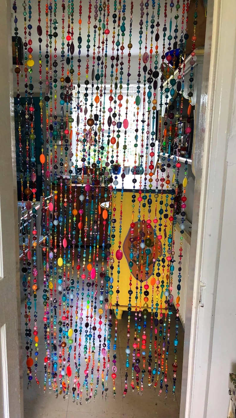 Pu&ograve; includere: Una tenda per porta fatta di fili di perline colorate. Le perline sono di varie forme e dimensioni, tra cui rotonde, ovali e oblunghe, nelle tonalit&agrave; di rosso, arancione, giallo, verde, blu e viola. La tenda &egrave; appesa davanti a un mobile giallo.