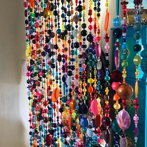 Pu&ograve; includere: Una vivace tenda di perline con fili di perline colorate di varie forme e dimensioni. Le perline sono nelle tonalit&agrave; di rosso, arancione, giallo, verde, blu, viola e rosa. La tenda &egrave; appesa davanti a un muro bianco.