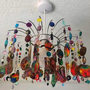 May include: A colourful chandelier with a metal frame and hanging glass beads, crystals, and painted metal ornaments. The chandelier has a white ceiling mount and a clear light bulb.