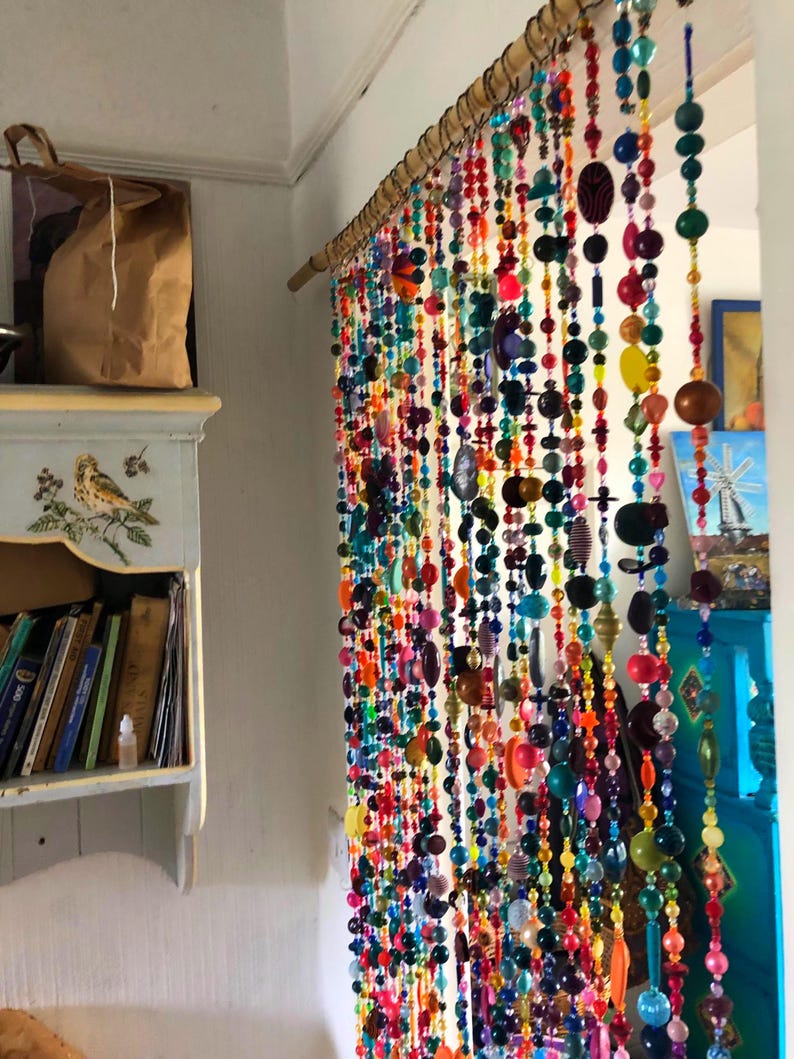 Pu&ograve; includere: Una colorata tenda di perline &egrave; appesa in una porta, con perline di varie forme e dimensioni. Le perline sono infilate in fili verticali. I colori includono rosso, arancione, giallo, verde, blu e viola, creando un elemento decorativo vivace.