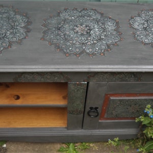 May include: A grey wooden cabinet with a decorative mandala design painted on the top. The cabinet has a single door and two shelves.