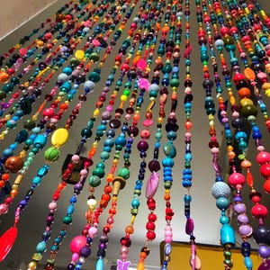 Pu&ograve; includere: Una vibrante tenda di perline multicolore &egrave; appesa al soffitto. La tenda &egrave; composta da numerosi fili, ciascuno adornato con perline in una vasta gamma di colori, tra cui rosso, arancione, giallo, verde, blu e viola. Le perline variano per dimensioni e forma.