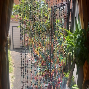 Pu&ograve; includere: Una tenda di perline colorate &egrave; appesa davanti a una finestra. Le perline, di varie forme e dimensioni, sono nei colori rosso, arancione, giallo, verde, blu e viola. Attraverso la tenda si vede un giardino.