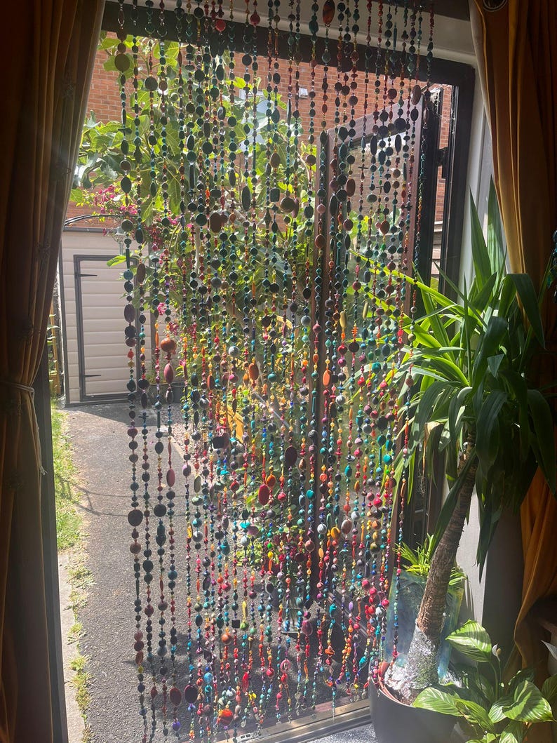 Pu&ograve; includere: Una tenda di perline colorate &egrave; appesa davanti a una finestra. Le perline, di varie forme e dimensioni, sono nei colori rosso, arancione, giallo, verde, blu e viola. Attraverso la tenda si vede un giardino.