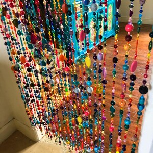 Pu&ograve; includere: Una vivace tenda di perline con fili di perline colorate di varie forme e dimensioni. Le perline sono nelle tonalit&agrave; di rosso, arancione, giallo, verde, blu e viola. La tenda &egrave; appesa davanti a un mobile turchese e un pavimento in legno.