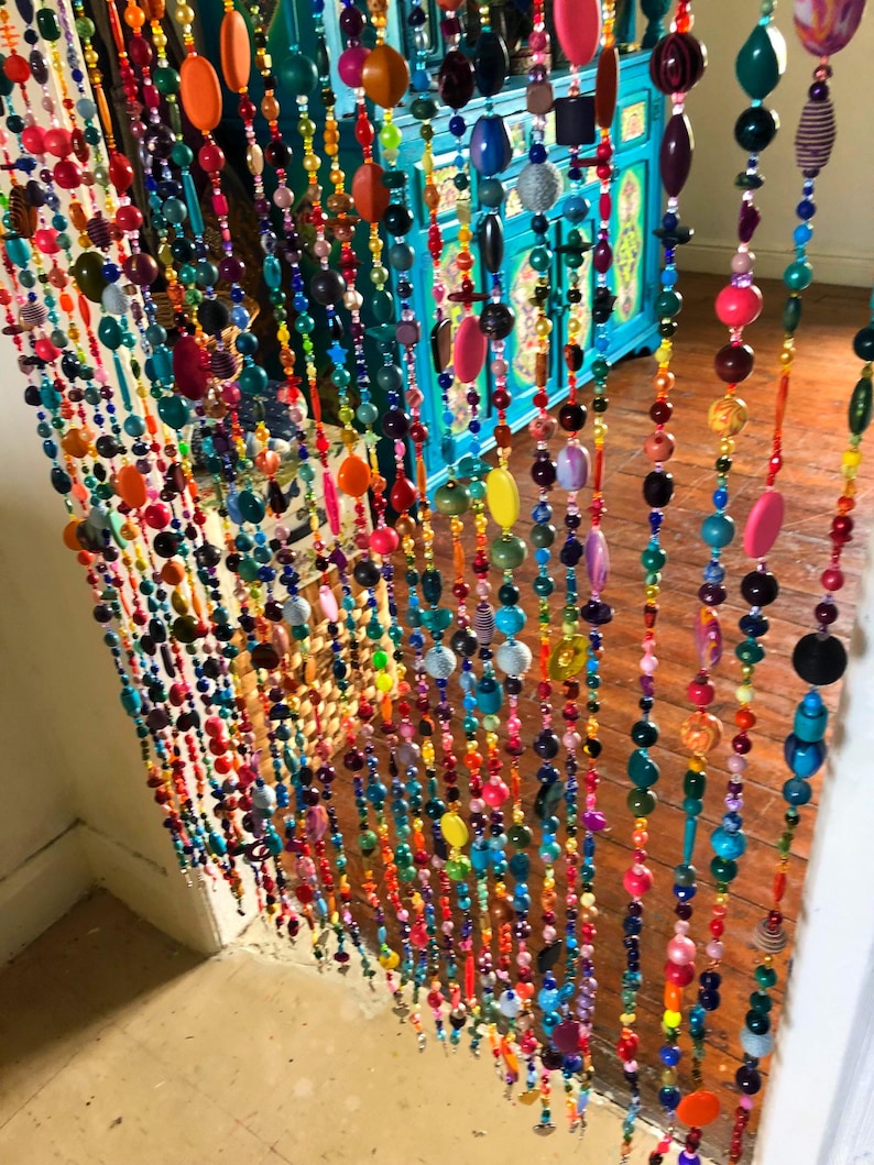 Pu&ograve; includere: Una vivace tenda di perline con fili di perline colorate di varie forme e dimensioni. Le perline sono nelle tonalit&agrave; di rosso, arancione, giallo, verde, blu e viola. La tenda &egrave; appesa davanti a un mobile turchese e un pavimento in legno.