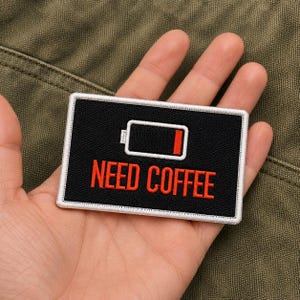 May include: Black rectangular patch with white border. The patch features a battery icon with a low charge and the words "NEED COFFEE" in red. The patch is held in a hand against a green background.