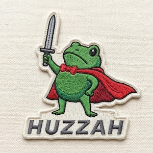 May include: Embroidered patch featuring a green frog wearing a red cape and bow tie, holding a sword. The patch has the word "Huzzah" embroidered in gray letters on a white background. The frog is the central focus of the patch.