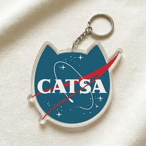 May include: Clear acrylic keychain with a cat-shaped design. The keychain features a blue circle with the text "CATSA" in white, a red swoosh, and white stars, resembling the NASA logo. Includes a silver keyring and chain.