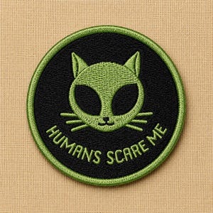 May include: Embroidered patch featuring a green alien cat face on a black circle, with the text "HUMANS SCARE ME" in green. The patch has a green border and is set against a tan fabric background.