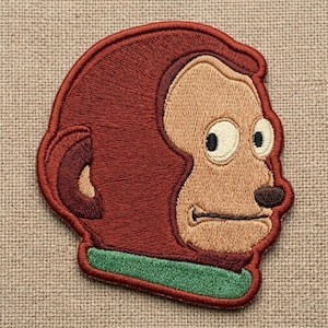 May include: Embroidered patch featuring a cartoon monkey head in profile. The patch has a brown outline and a reddish-brown head with a tan face. The monkey has large, wide eyes, a small brown nose, and a green base.