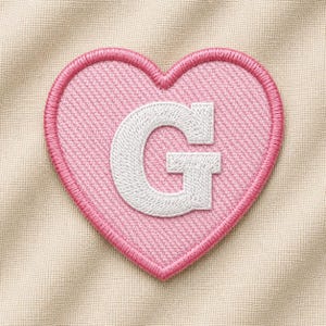 May include: A pink heart-shaped embroidered patch with a white letter "G" in the center. The patch has a pink border and is set against a beige fabric background. The design is simple and clean, suitable for various applications.