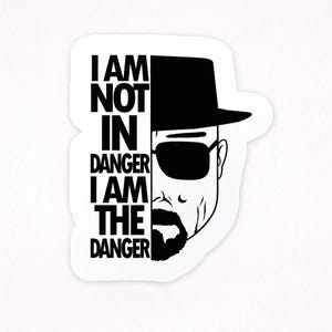 May include: A white sticker featuring a black and white graphic of a man wearing a hat and sunglasses. The text "I AM NOT IN DANGER I AM THE DANGER" is printed in bold, black letters.