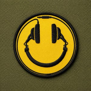May include: Embroidered patch featuring a yellow smiley face with black headphones forming the eyes and a smile. The patch has a black border and is set against a green background. The headphones are connected by a black cable.