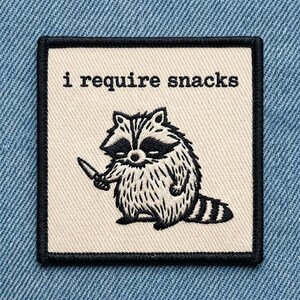 May include: Square patch with a black border and the text "i require snacks." The patch features a black and white cartoon raccoon holding a knife. The patch is on a light beige background.