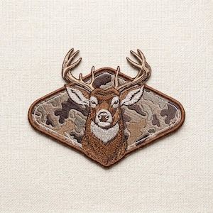 May include: Embroidered patch featuring a deer head with large antlers. The deer is brown with white accents, set against a camouflage background within a brown diamond shape. The patch is designed for apparel or accessories.