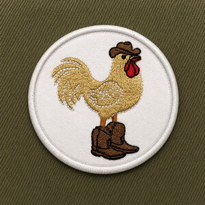 May include: Embroidered patch featuring a cartoon rooster wearing a cowboy hat and boots. The rooster is gold with a red wattle and comb, set against a white circular background with a white border. The background is olive green.