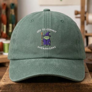 May include: A green baseball cap with the text "OFF TO COMMIT SHENANIGANS" and an embroidered frog wizard design. The frog is wearing a purple robe and hat, holding a staff. The cap has a curved brim and a button on top.