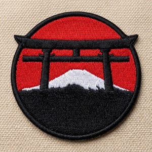 May include: Embroidered patch featuring a Japanese landscape. The design includes a red sun, a black torii gate, a white mountain, and a black mountain base, all set within a black circular border. The patch is on a tan fabric background.