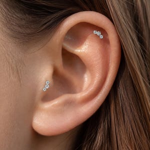 Könnte beinhalten: Zwei silberfarbene Ohrpiercings mit einem Trio aus kleinen, klaren, rund geschliffenen Steinen, die in einer geschwungenen Linie angeordnet sind. Ein Schmuckstück befindet sich am Tragus, das andere am oberen Knorpel des Ohrs.