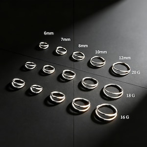 Puede incluir: Una colección de pendientes de aro dobles plateados en varios tamaños, que van desde 6 mm hasta 12 mm, y calibres de 16G a 20G. Los pendientes se muestran sobre una superficie oscura, con etiquetas de tamaño.