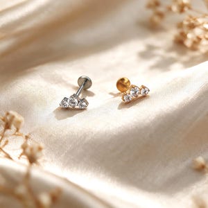 May include: Two flat back stud earrings, one in silver-tone metal and one in gold-tone metal. Each earring features a curved bar design set with three clear, round-cut stones arranged in a cluster. The posts have a circular flat back closure.