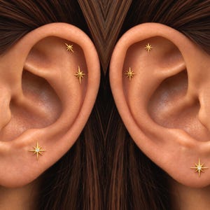 Könnte beinhalten: Ein Set aus goldfarbenen, sternförmigen Ohrsteckern mit einem vielzackigen Design. Jeder Ohrstecker ist mit einem zentralen, klaren Stein besetzt. Die Ohrringe sind am Ohrläppchen und im Knorpelbereich platziert.