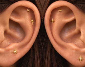 May include: A set of gold star-shaped stud earrings featuring a central clear stone. Each ear displays three individual star studs positioned along the cartilage and the lobe, creating a celestial-inspired arrangement of jewellery.