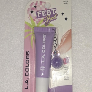 May include: L.A. Colors candy lipgloss in a light purple squeeze tube with a matching purple pom-pom keychain attached. The packaging features the text FEST Best, L.A. Colors, and 0.34 FL. OZ. (10 mL) on a card with a disco ball and palm tree graphic.