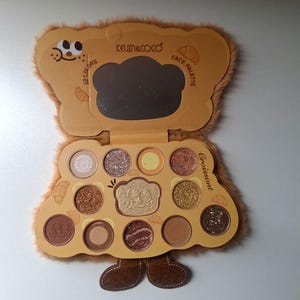 K&ouml;nnte beinhalten: Braune Lidschatten-Palette in B&auml;renform mit 12 Farben, einem Spiegel und dem Text "KEVIN&COCO" und "FACE PALETTE". Das Geh&auml;use hat eine flauschige Textur und ein Cartoon-B&auml;r-Design. Das Wort "Croissant" ist ebenfalls sichtbar.