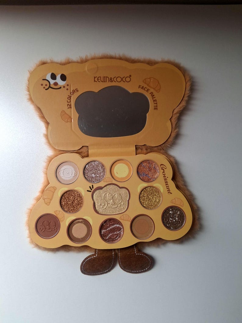 K&ouml;nnte beinhalten: Braune Lidschatten-Palette in B&auml;renform mit 12 Farben, einem Spiegel und dem Text "KEVIN&COCO" und "FACE PALETTE". Das Geh&auml;use hat eine flauschige Textur und ein Cartoon-B&auml;r-Design. Das Wort "Croissant" ist ebenfalls sichtbar.