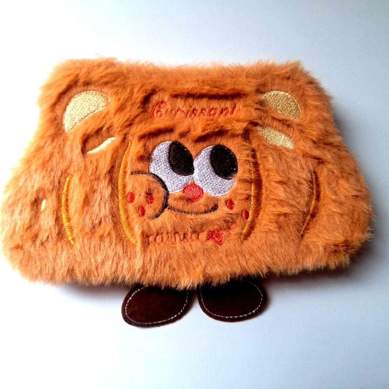 K&ouml;nnte beinhalten: Ein orange-braunes Federm&auml;ppchen aus Pl&uuml;sch in Form eines Cartoon-Croissants. Es hat gestickte Details, darunter ein l&auml;chelndes Gesicht, das Wort "Croissant" und zwei braune F&uuml;&szlig;e. Das M&auml;ppchen hat eine weiche, pelzige Textur.